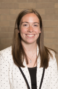 Hope Bibens, a woman with long, straight brown hair, smiles at the camera. She wears a white blazer with black polka dots and black trim and a silver key necklace.